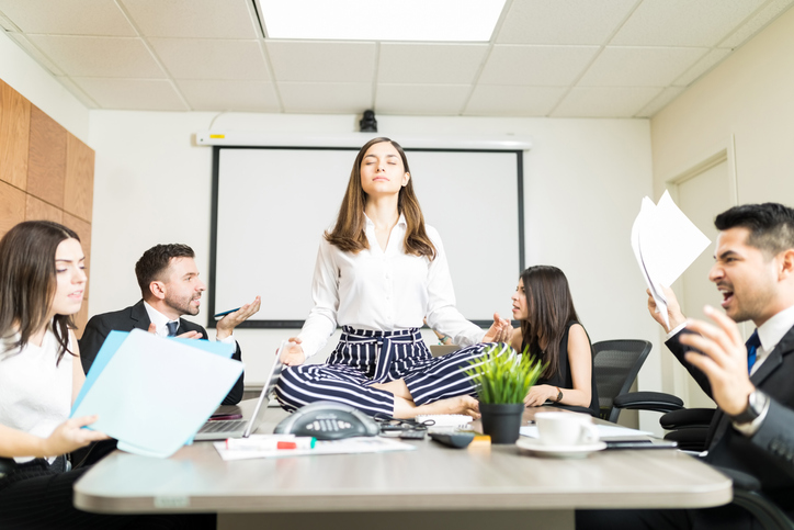 Woman sitting on a conference table meditating while others compete for her attention.